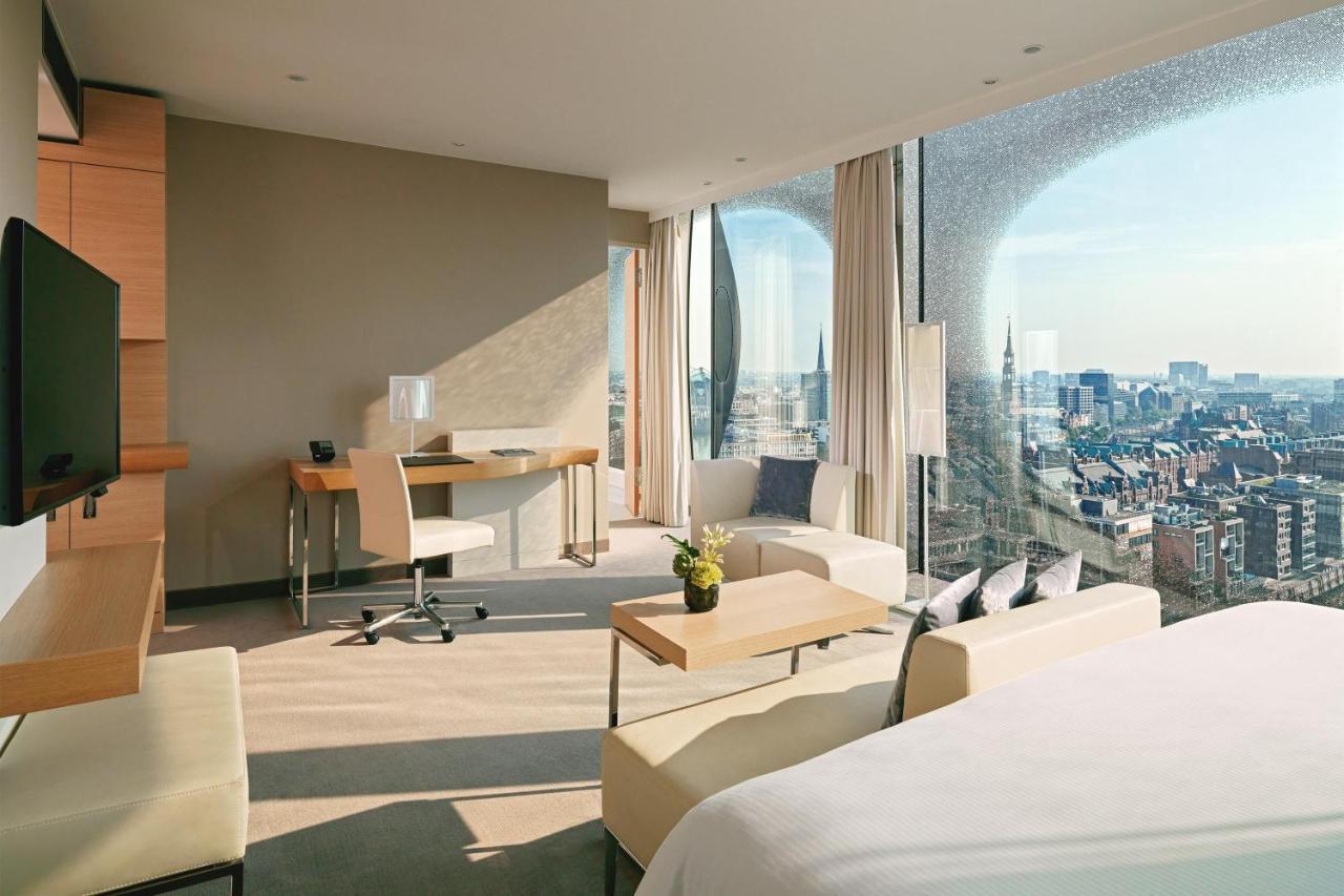 The Westin Hamburg Elbphilharmonie Hotel Exterior photo The photo depicts a modern hotel room featuring a contemporary design. The space includes a large window that offers a panoramic view of the city beyond. Inside, there are stylish furniture elements such as a small dining table and chairs, a desk wit