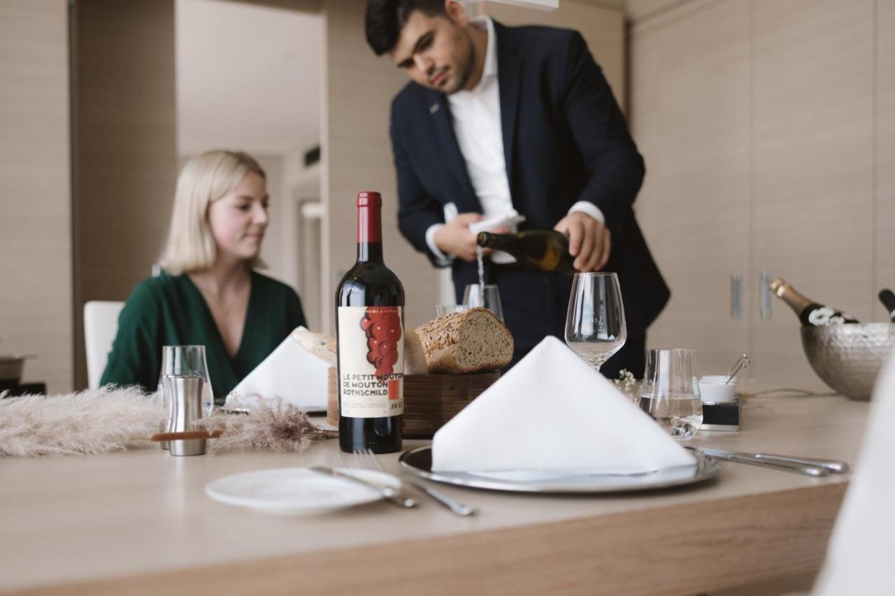 The Westin Hamburg Elbphilharmonie Hotel Exterior photo The photo shows a dining scene featuring a well-dressed man pouring wine into a glass while a woman sits at the table. The table is elegantly set with a folded napkin, wine glasses, and a bottle of red wine placed prominently. There is also a loaf of