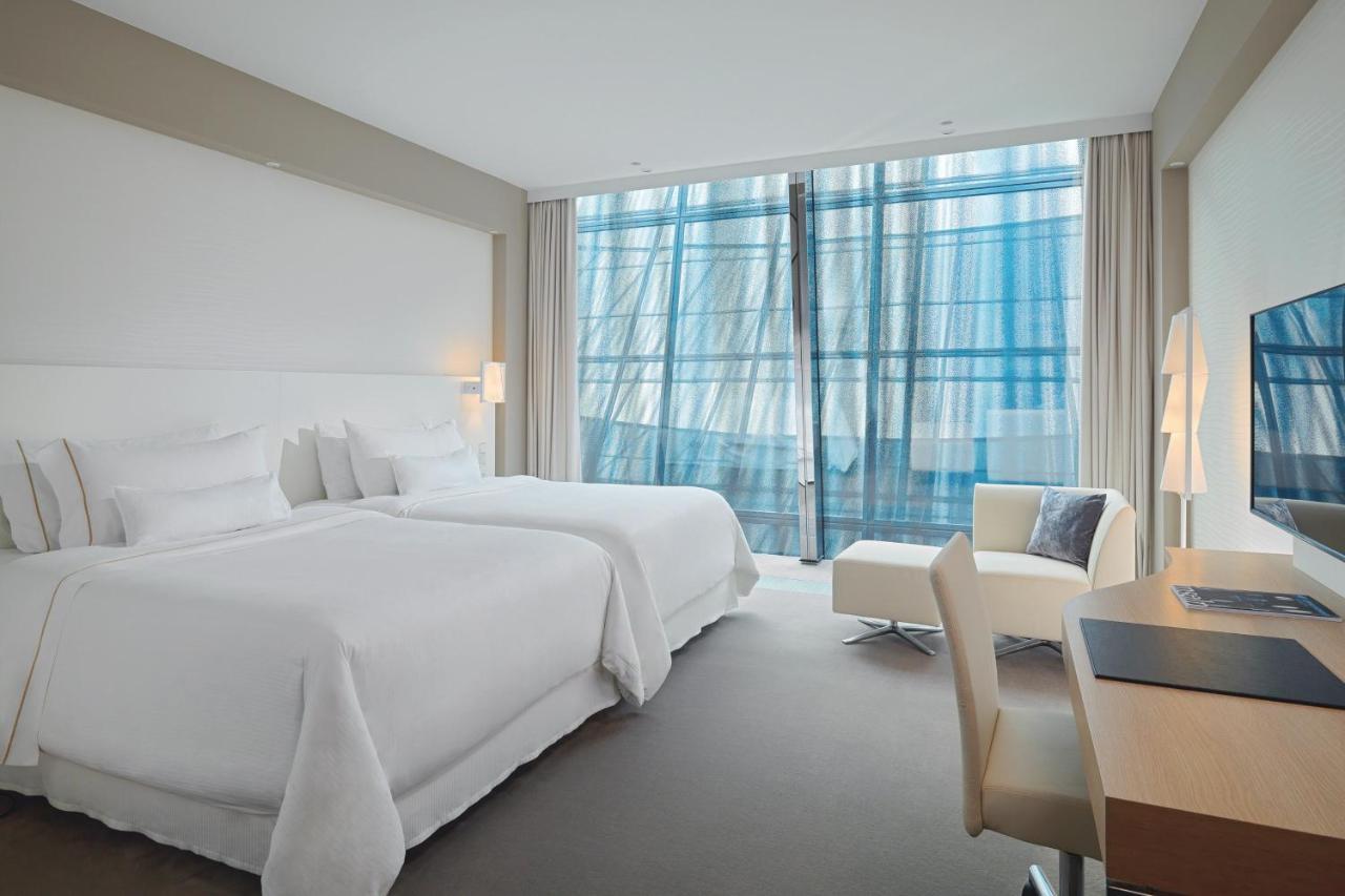 The Westin Hamburg Elbphilharmonie Hotel Exterior photo The photo shows a modern hotel room featuring two white beds with plush bedding. There is a large window with sheer curtains that allows natural light to filter in, offering a view outside. The room has neutral-colored walls and carpeting, creating a