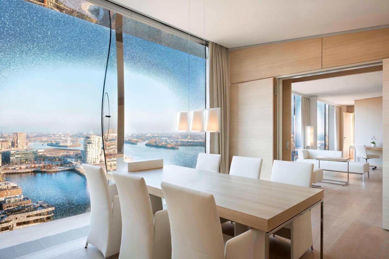 The Westin Hamburg Elbphilharmonie Hotel Exterior photo The photo shows a modern dining area with a large, light-colored table surrounded by white chairs. The space features a panoramic window that offers a stunning view of a body of water and a cityscape. The decor is minimalistic and elegant, with smoot