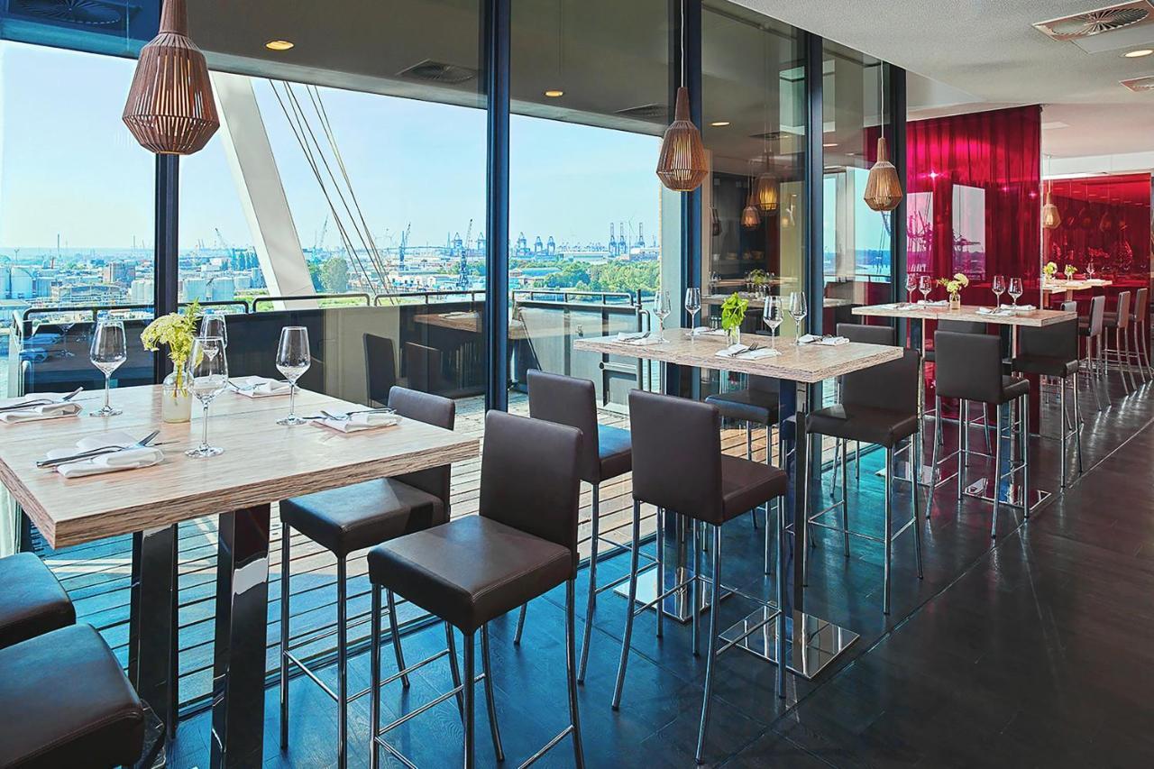 The Westin Hamburg Elbphilharmonie Hotel Exterior photo The photo depicts a modern restaurant interior featuring a spacious dining area with large windows that provide a view of the outside. There are several tall tables with a light-colored surface and metallic bases, accompanied by dark, sleek stools. T