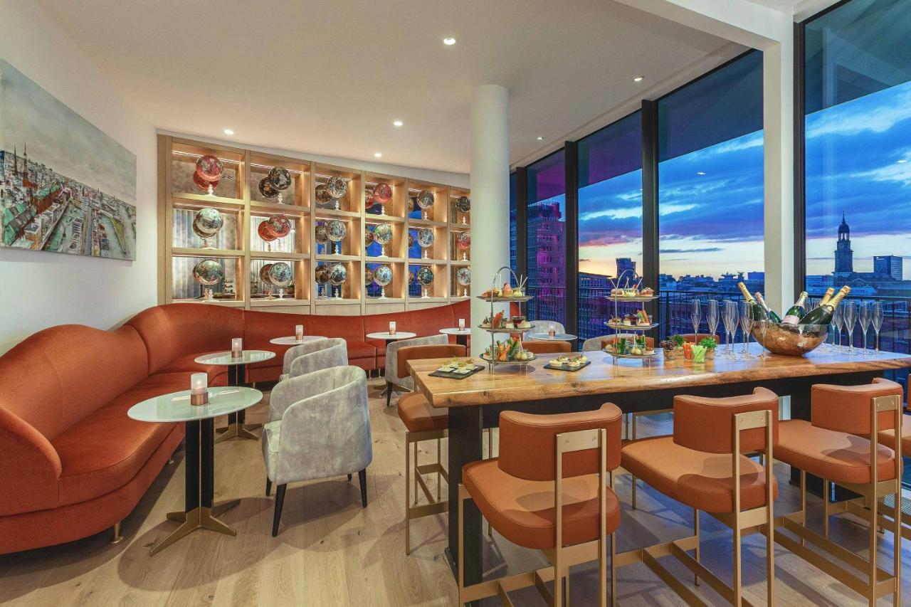 The Westin Hamburg Elbphilharmonie Hotel Exterior photo The photo shows a modern and stylish restaurant or café interior. It features a combination of comfortable seating, including a curved, orange sofa and gray chairs. The tables are round with light-colored tops, and there are several tiered stands dis