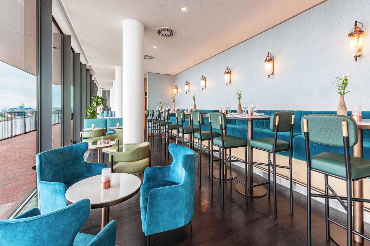 The Westin Hamburg Elbphilharmonie Hotel Exterior photo The photo shows a modern and stylish restaurant or café interior. It features a long row of high-backed bar stools at a counter, complemented by plush seating in the form of rounded chairs and small tables arranged throughout the space. The color sch