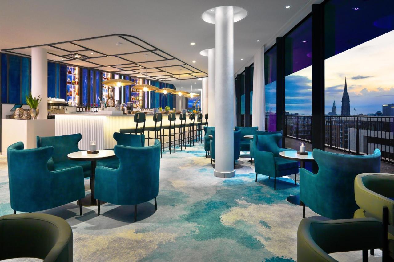 The Westin Hamburg Elbphilharmonie Hotel Exterior photo The photo shows a modern lounge or bar area with a stylish design. It features plush turquoise chairs and small tables arranged around the space. In the background, there's a long bar with seating, illuminated by soft lighting. Large windows provide 