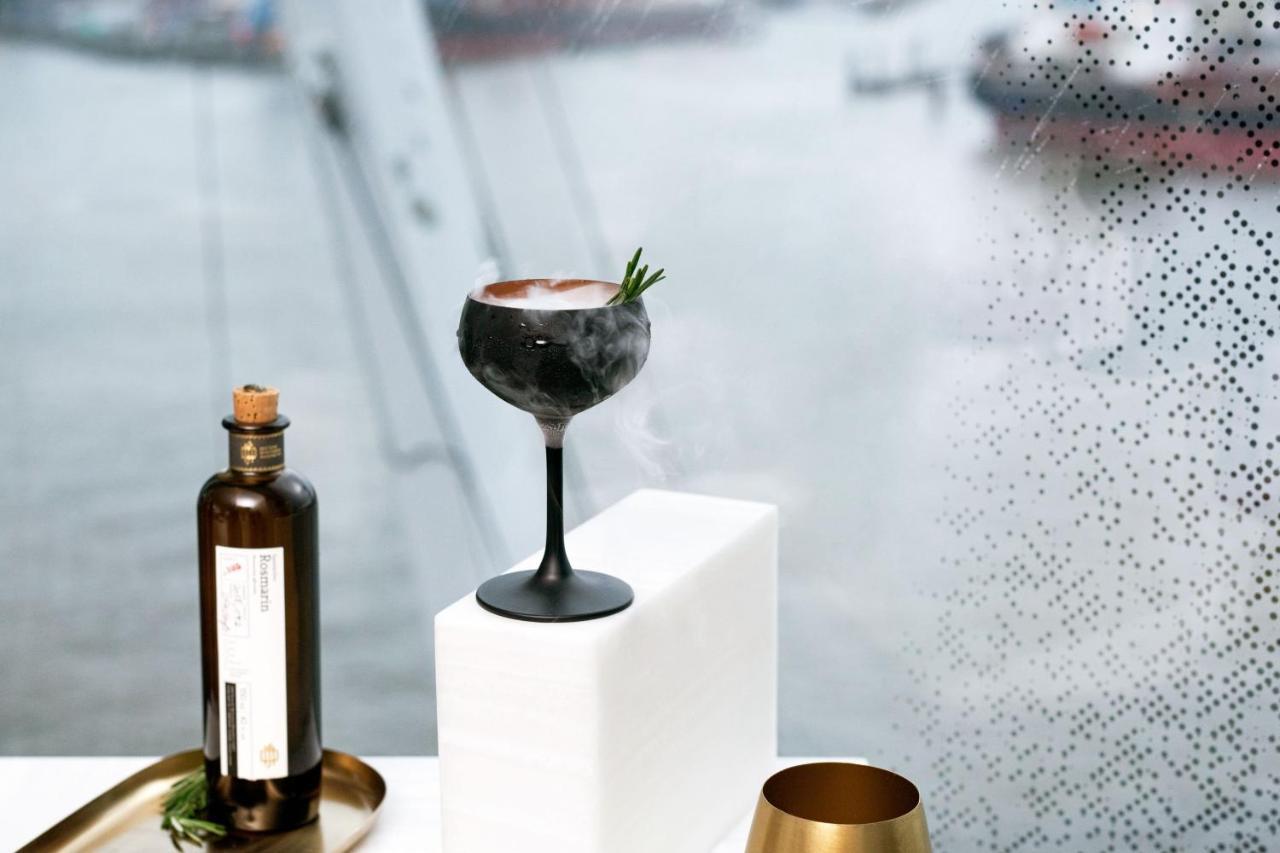 The Westin Hamburg Elbphilharmonie Hotel Exterior photo The photo features a stylish cocktail elegantly presented on a white pedestal. The cocktail is served in a sophisticated black stem glass, topped with a garnish of rosemary. Beside it, there is a dark glass bottle, likely containing a spirit or mixer