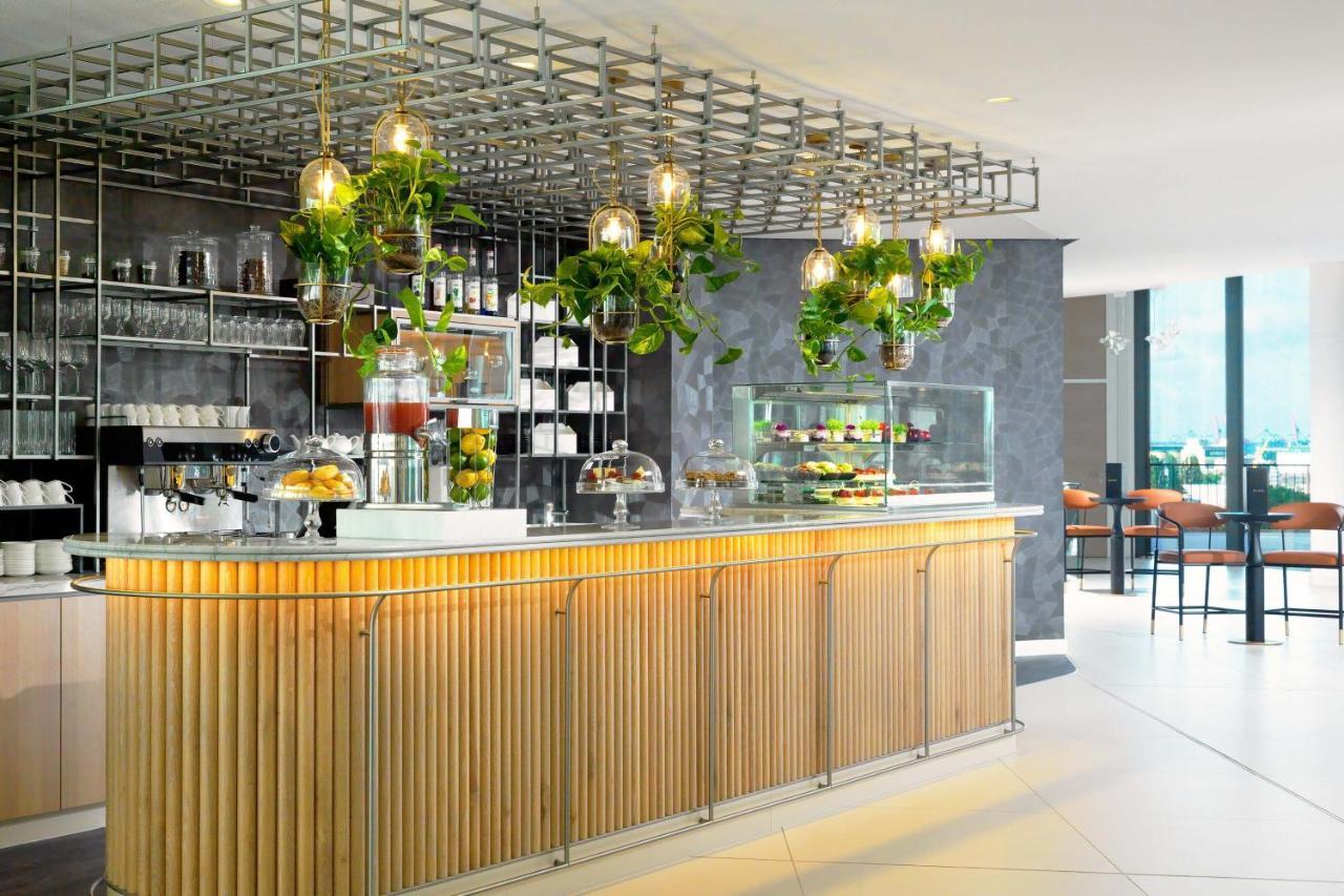 The Westin Hamburg Elbphilharmonie Hotel Exterior photo The photo shows a modern café or bar setting with a sleek wooden counter that features vertical slats. Above the counter, there is a grid-like framework hanging from the ceiling, adorned with various green plants that add a touch of nature to the int