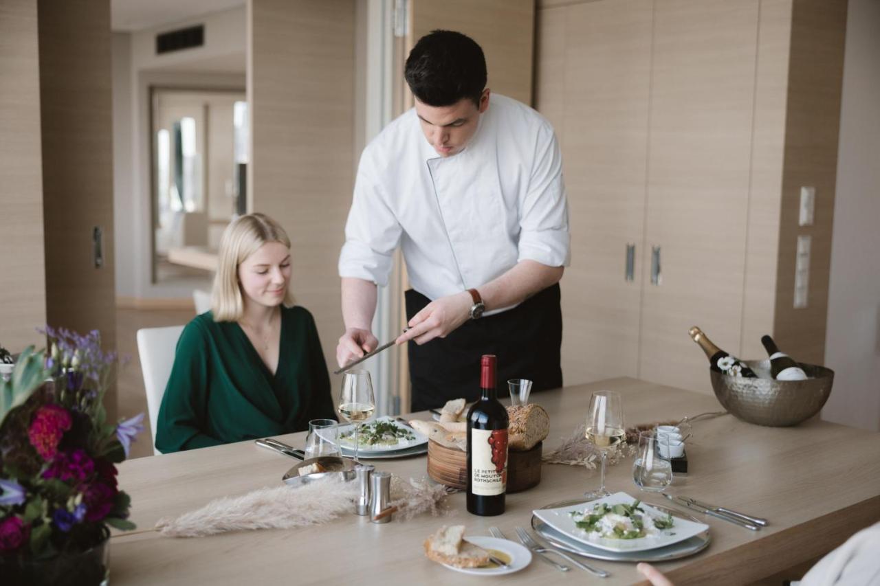 The Westin Hamburg Elbphilharmonie Hotel Exterior photo The photo depicts a dining scene in a modern setting. A man, dressed in a chef's attire, is serving food to a woman seated at the table. The woman is wearing a green outfit and appears to be enjoying the meal. The table is elegantly set with plates o