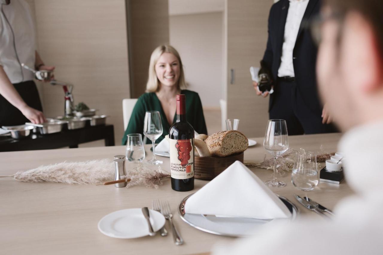 The Westin Hamburg Elbphilharmonie Hotel Exterior photo The image shows a dining setting with a woman smiling at the table. She is seated in front of a bottle of red wine that has a distinctive label. There is also bread and a neatly arranged table setting, including plates and glasses. In the background,