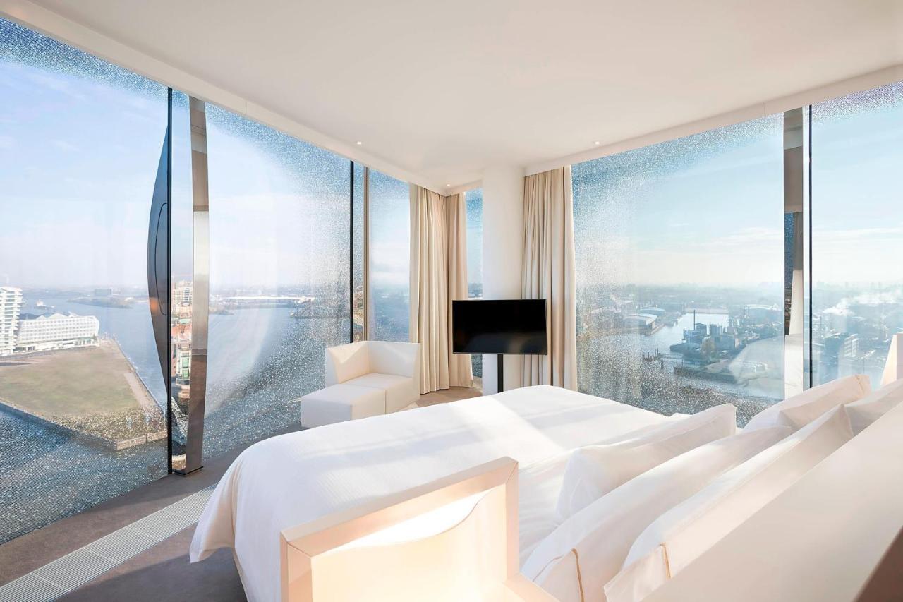 The Westin Hamburg Elbphilharmonie Hotel Exterior photo The photo shows a modern hotel room featuring a large bed with white linens and a minimalist design. It has expansive floor-to-ceiling windows that offer a panoramic view of the landscape outside, likely a city or waterfront setting. The room is deco