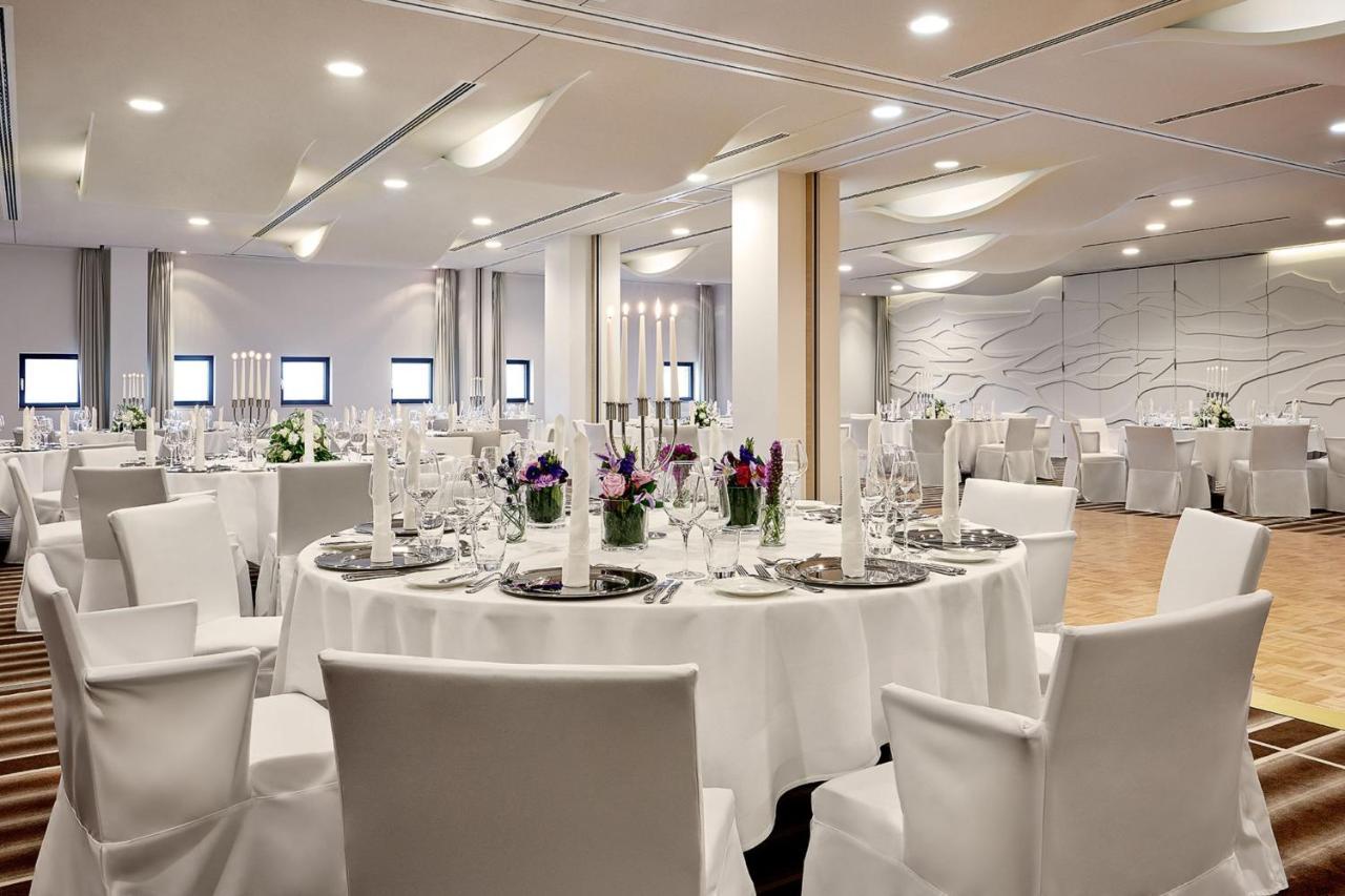 The Westin Hamburg Elbphilharmonie Hotel Exterior photo The photo depicts a modern banquet or event hall setup. It features round tables covered with white tablecloths, each set with elegant dishware and glassware. The tables are adorned with floral centerpieces, adding a touch of color, and there are tal