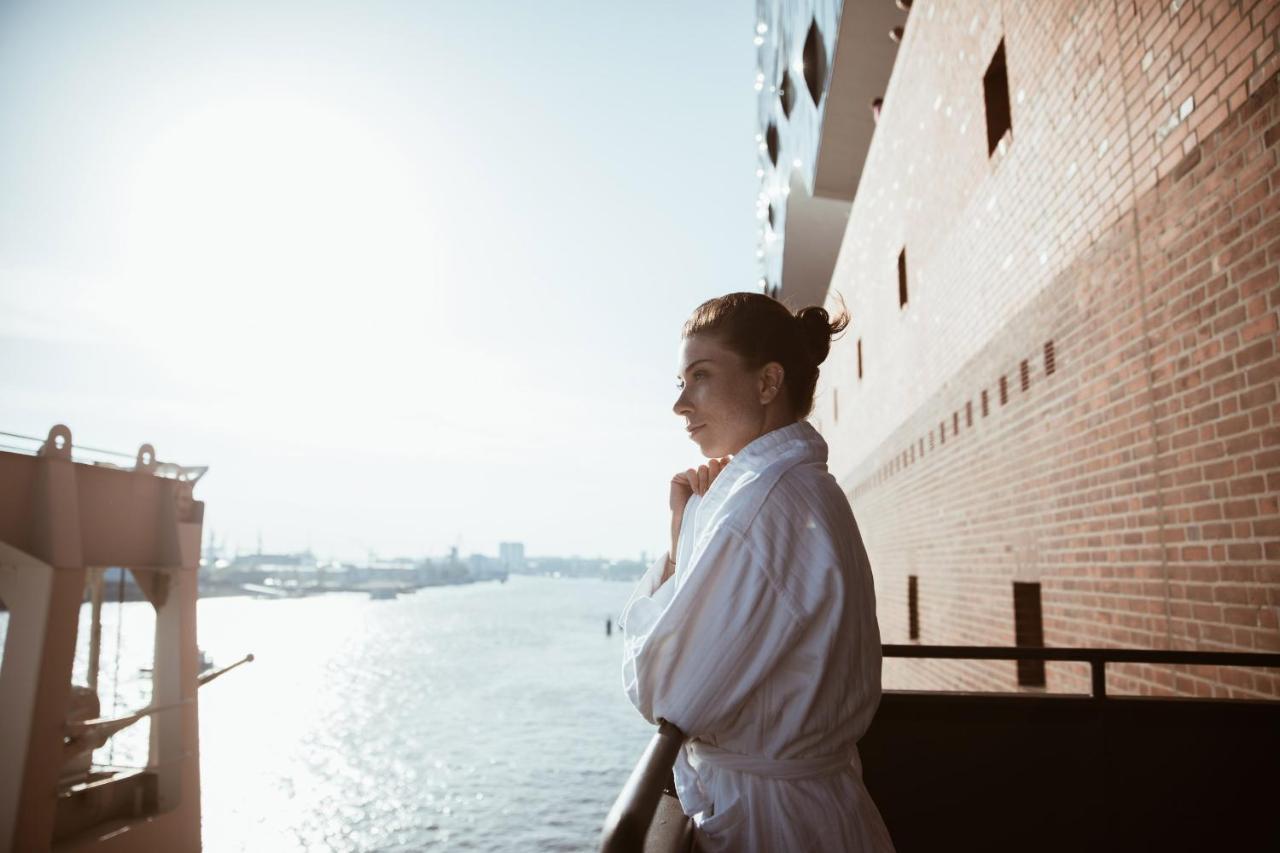 The Westin Hamburg Elbphilharmonie Hotel Exterior photo The photo depicts a person standing on a balcony, looking out over a body of water. The individual is wearing a white bathrobe, and their hair is styled in a bun. The background features a scenic view with sunlight illuminating the scene, creating a 