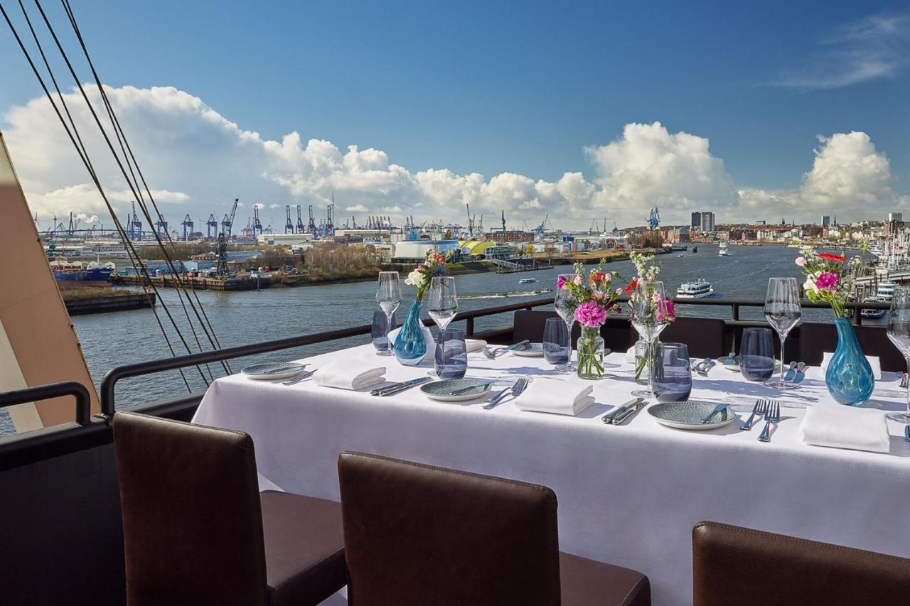 The Westin Hamburg Elbphilharmonie Hotel Exterior photo The photo shows a beautifully set dining table on a rooftop or terrace with a view of a body of water. The table is adorned with a white tablecloth, elegant tableware, and multiple vases filled with colorful flowers. In the background, there are boat