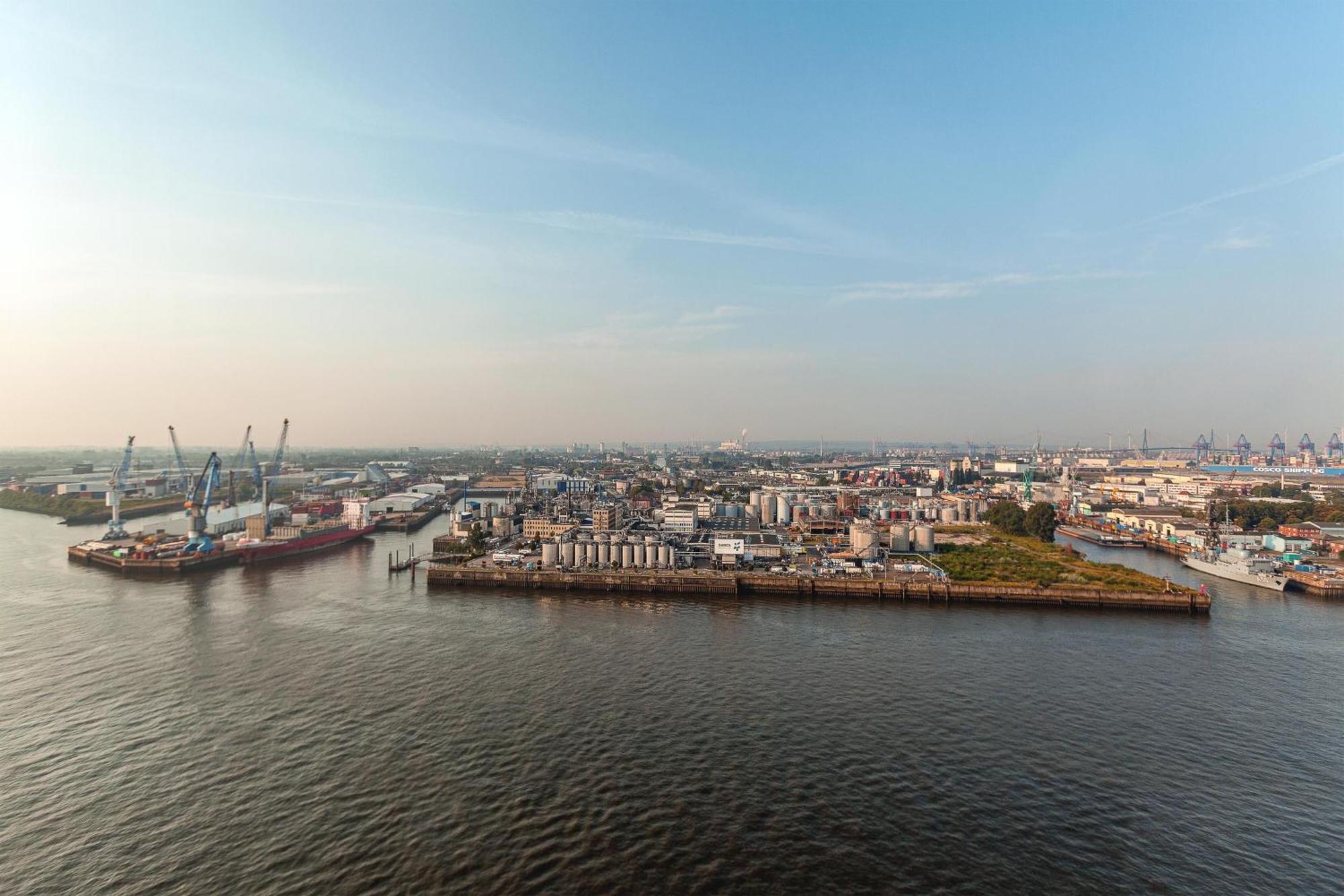 The Westin Hamburg Elbphilharmonie Hotel Exterior photo The photo shows a coastal area with a large body of water in the foreground. To the left, there are industrial structures like cranes and cargo ships, indicating an active port or shipping area. In the background, there are various buildings, which a