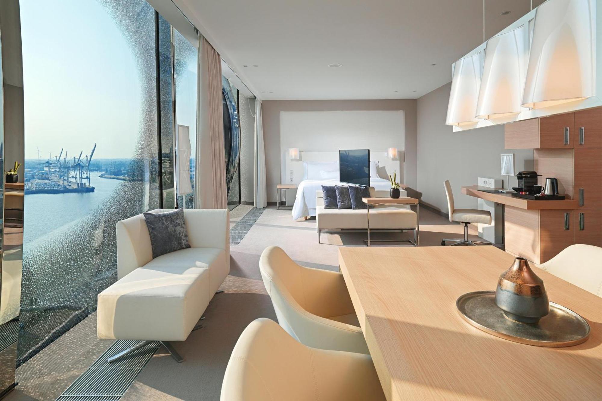 The Westin Hamburg Elbphilharmonie Hotel Exterior photo The photo shows a modern hotel room with a spacious layout. On one side, there are large floor-to-ceiling windows that offer a view of the outside, likely showcasing a waterfront or cityscape. Inside, there's a bed with a minimalist design, accompani