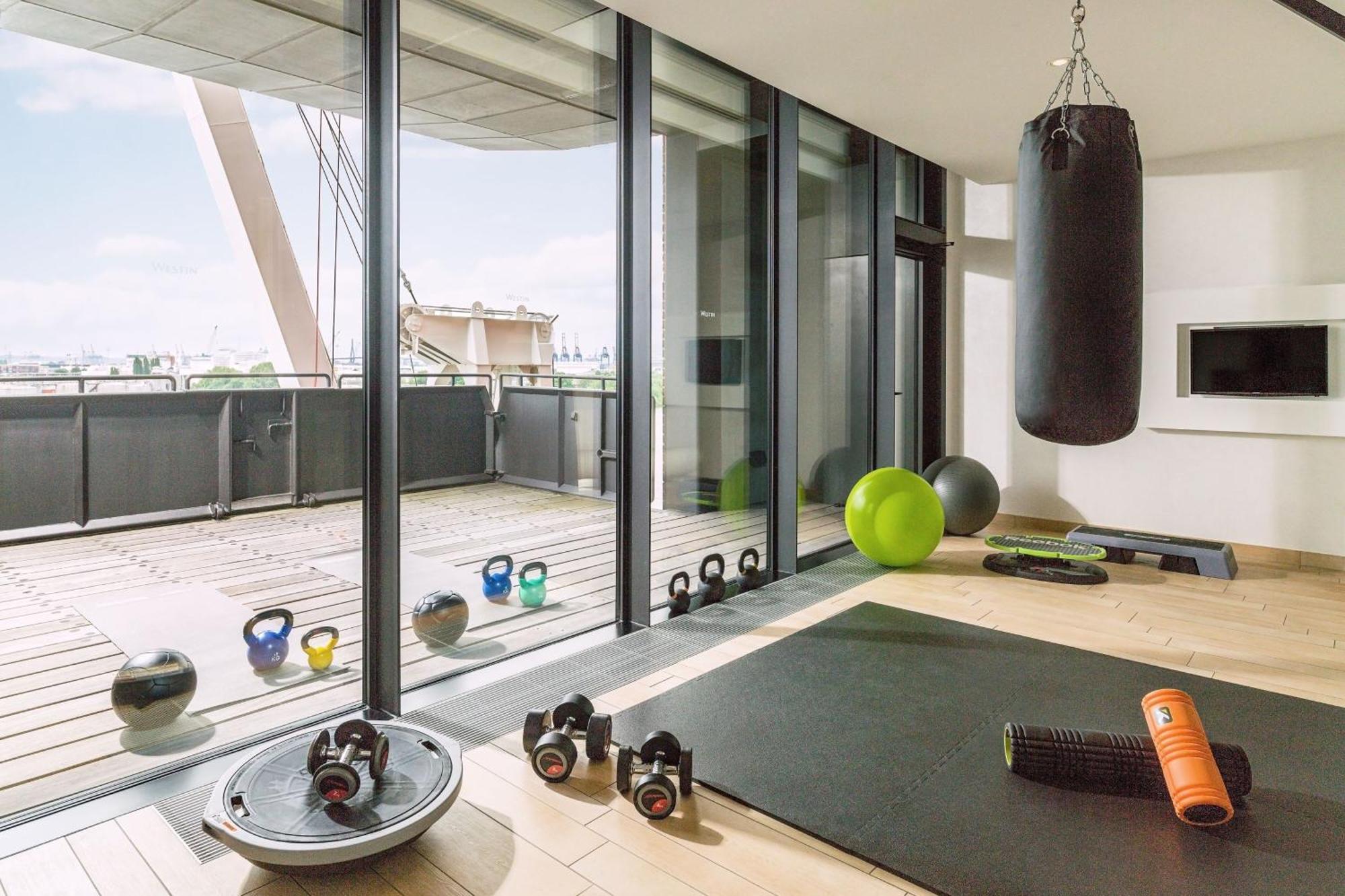 The Westin Hamburg Elbphilharmonie Hotel Exterior photo The photo shows a modern home gym with large glass windows that provide a view to the outside. Inside, there are various fitness equipment items, including a heavy punching bag hanging from the ceiling, kettlebells, a stability ball, an exercise mat,
